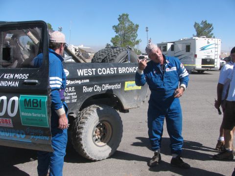 1990 Ford Bronco - Two men in blue racing suits talk beside the number 300 West Coast Broncos race truck with RVs and trees behind.