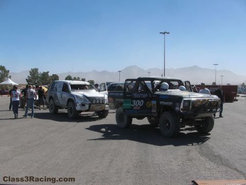1990 Ford Bronco - silver off-road SUV and the black number 300 race truck stage side by side in a dusty paved lot under mountains.
