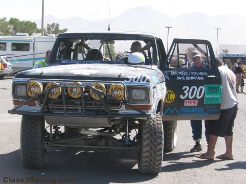 1990 Ford Bronco - Front view of the black number 300 race truck with a yellow HID light bar and open door, people gathered beside it.