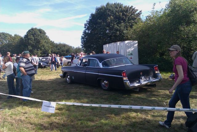 Black 1955 Chrysler New Yorker sedan parked on grass at a car show, people in background