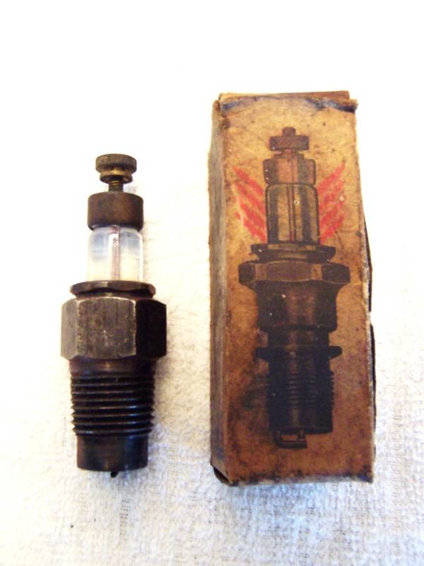 Antique brass spark plug with glass window beside its worn illustrated cardboard box