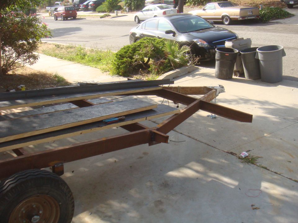 Trailer frame with wood planks, parked on driveway. Cars and trash cans in background