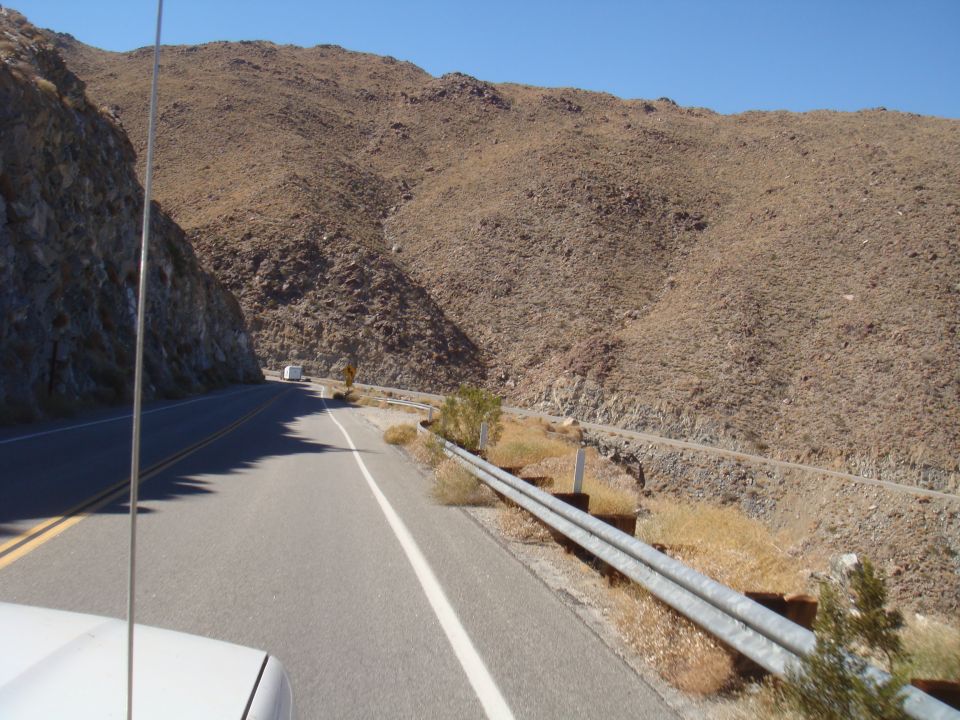 Driving on a two-lane road through a rocky, mountainous desert landscape under a clear blue sky. A white truck is visible far ahead