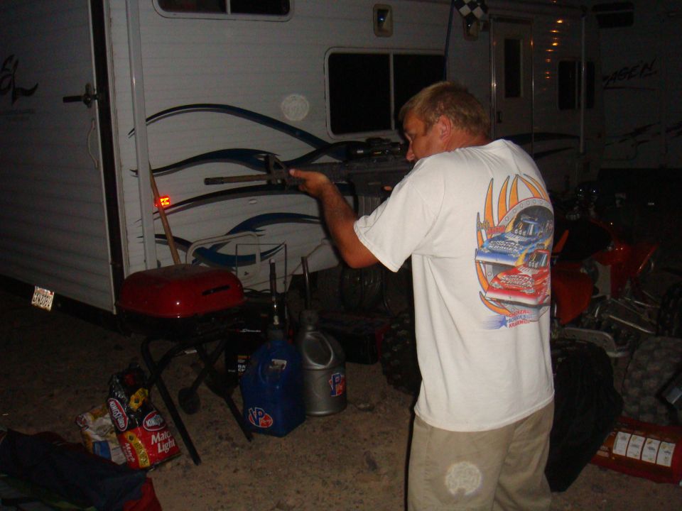 Man with blonde hair, wearing a white t-shirt with a racing graphic, holds a tool near a white trailer. A red ATV is partially visible