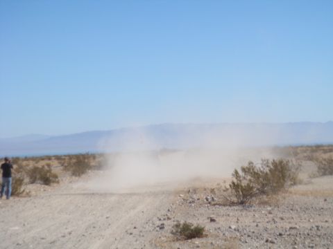 1992 Ford Bronco - A vehicle speeds across a desert landscape, leaving a trail of dust in its wake under a clear blue sky.