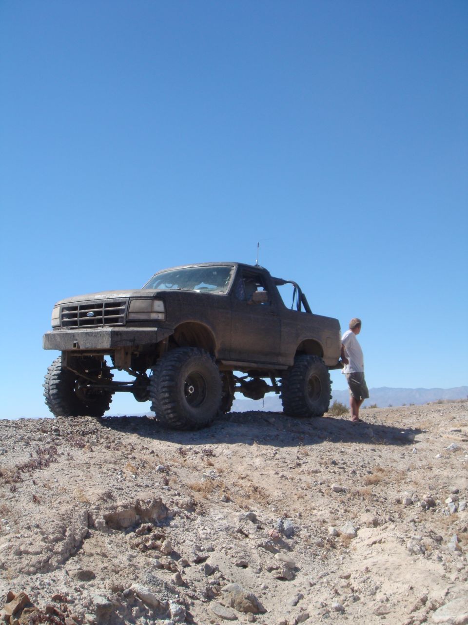 Dark 1992 Ford Bronco with large tires and roll cage, parked on a dirt hill under a clear blue sky