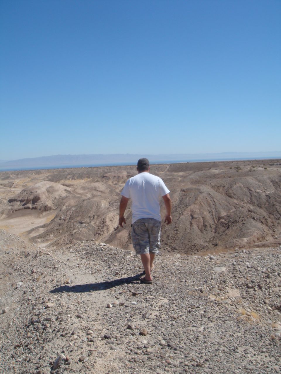 Man walks away from camera into a barren desert with mountains in the distance under a clear blue sky
