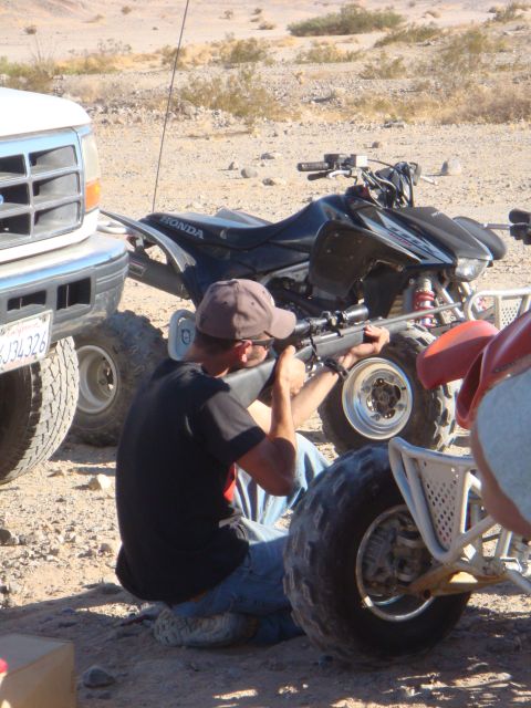 1992 Ford Bronco - A person aims a rifle near ATVs and a truck in a desert landscape.