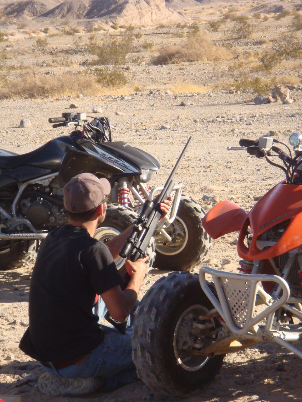 Person kneels with rifle between two ATVs in a desert landscape