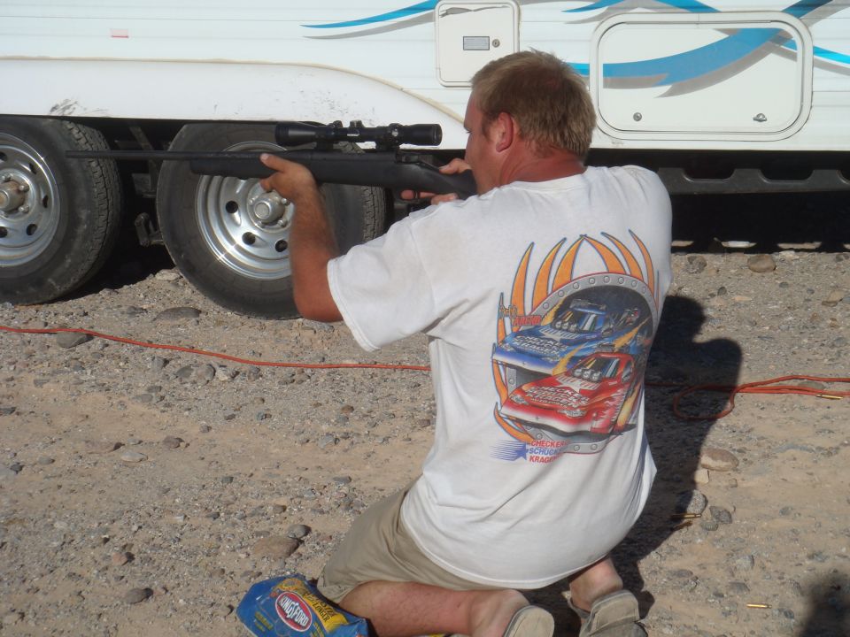 Man in white t-shirt aims rifle at the side of a white trailer with blue graphics