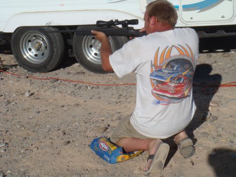 1992 Ford Bronco - Man aims rifle at trailer wheels while kneeling on the ground.