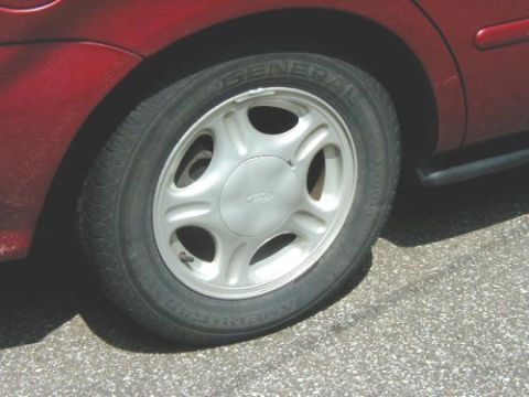 1997 Ford Taurus - Close-up of a red vehicle's rear wheel and fender, a five-spoke alloy wheel with a Ford emblem.