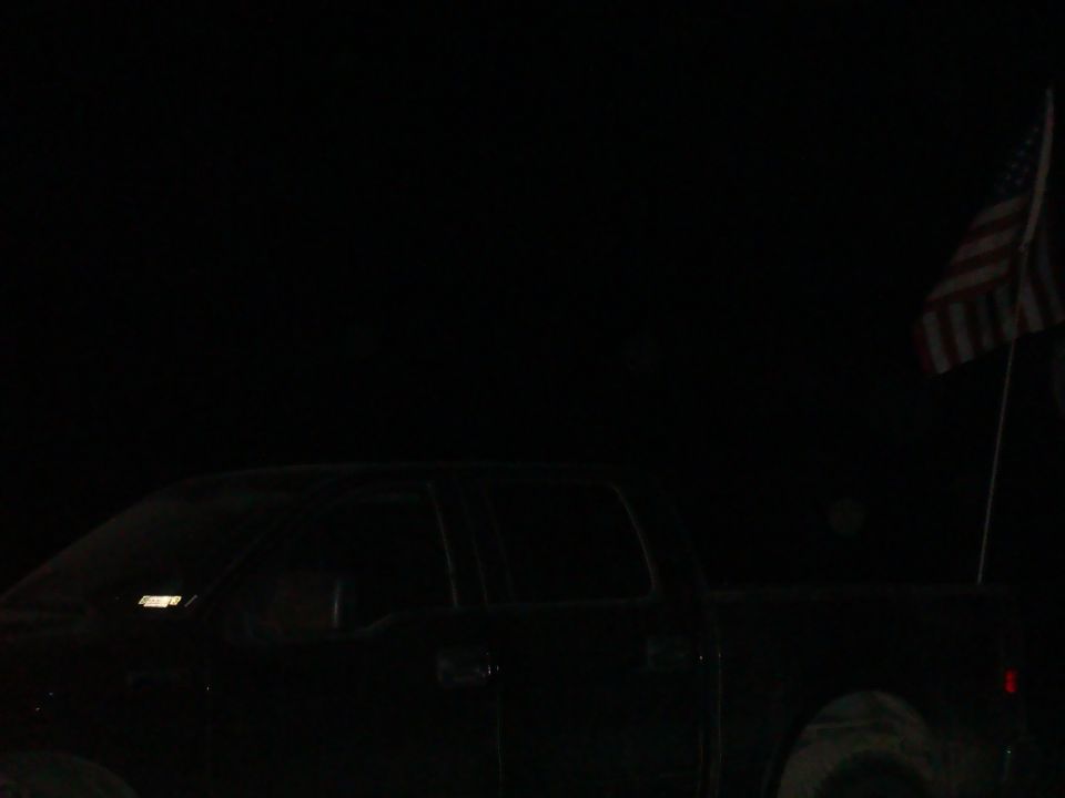 Dark pickup truck side profile, American flag on pole in background