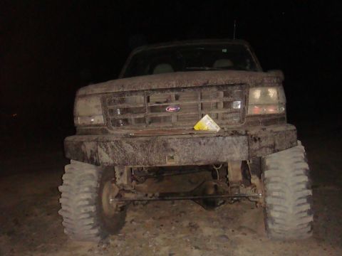 1992 Ford Bronco - A muddy 1992 Ford Bronco is seen from the front at night, showing its large tires and custom bumper.