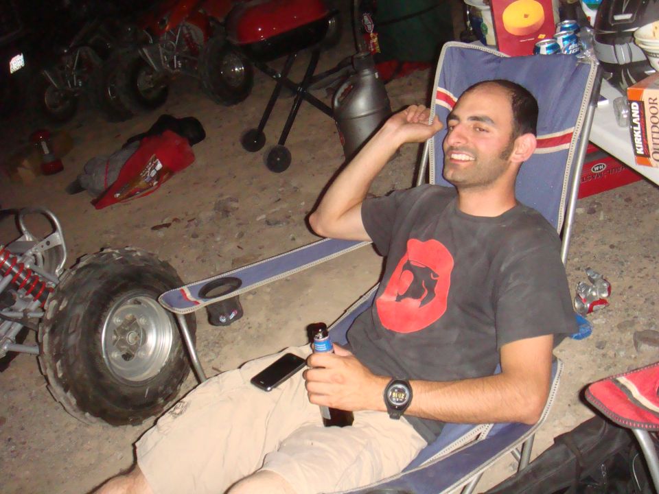 Guy chilling in a lawn chair with a beer, ATVs and a grill behind him