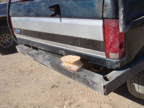 1992 Ford Bronco - A sandwich rests on the bumper of a dark green truck, which has a cracked taillight and a silver and black stripe on its tailgate.
