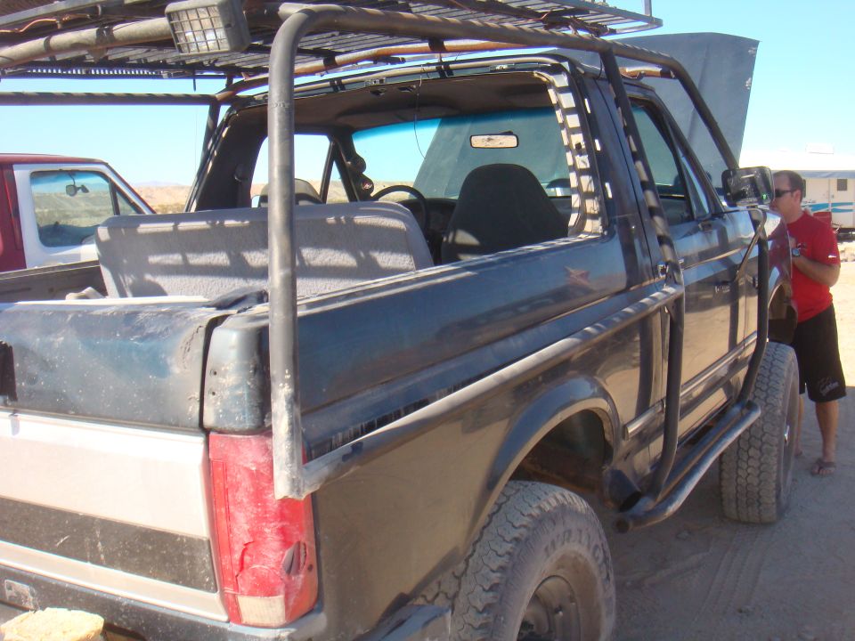 1992 Ford Bronco with roll cage, roof rack, and aftermarket wheels. Another vehicle is partially visible to the left. A person stands by the open hood