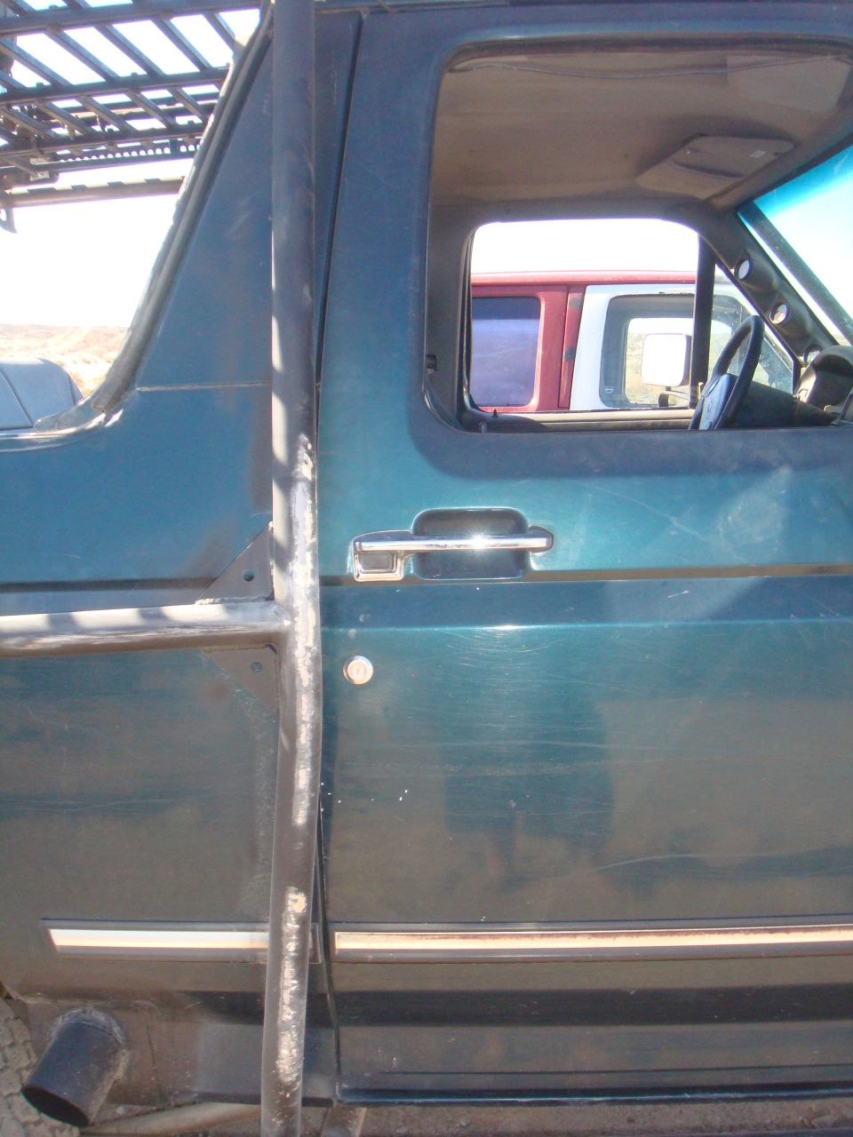 Side view of a 1992 Ford Bronco with a roof rack and roll cage. Another vehicle is visible through the window