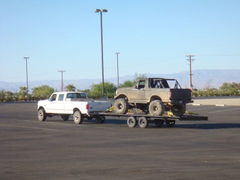 1992 Ford Bronco - A lifted 1992 Ford Bronco is transported on a trailer behind a white Ford F-Series truck in a parking lot.