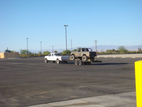 1992 Ford Bronco - Ford F-150 tows a lifted Ford Bronco on a trailer through a parking lot under a clear blue sky.