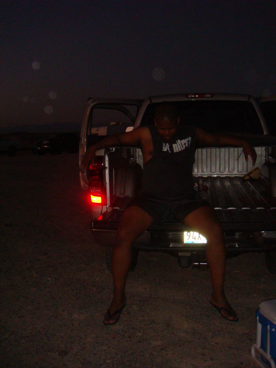 Man in shorts and t-shirt sits on the tailgate of a pickup truck at night. The truck's taillight is illuminated