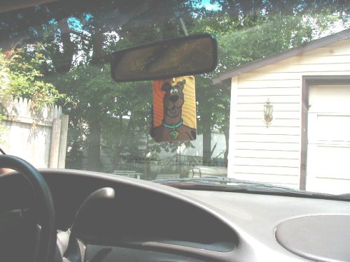 Car interior looking out windshield, shows dashboard, rearview mirror, and a Scooby-Doo air freshener hanging from it. A garage is visible outside