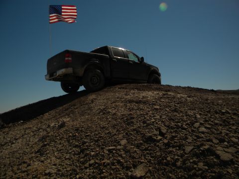 1992 Ford Bronco - A dark pickup truck is parked on a dirt hill under a clear blue sky with an American flag waving.