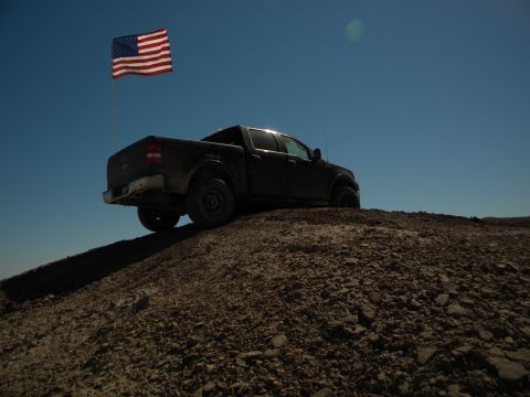 1992 Ford Bronco - A dark pickup truck is parked on a rocky incline, with an American flag waving in the clear blue sky above.