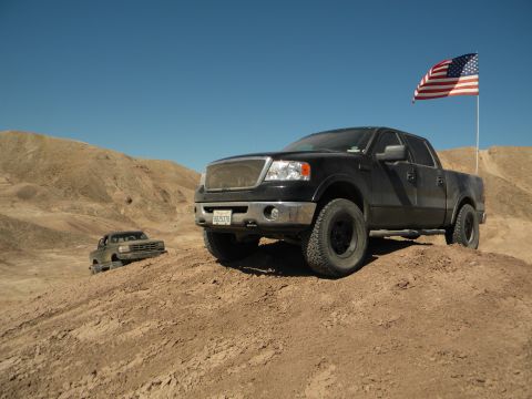 1992 Ford Bronco - A black pickup truck flies an American flag while parked on a dirt hill in a desert landscape, with another vehicle visible in the backgrou…
