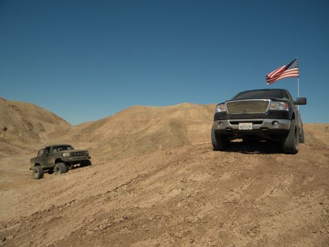 1992 Ford Bronco - Two trucks navigate a dusty desert terrain under a clear blue sky, with one truck flying an American flag.