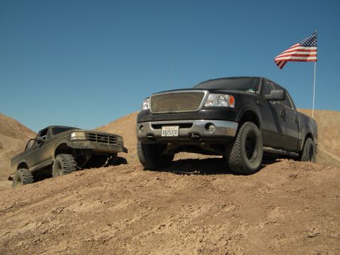 1992 Ford Bronco - Two trucks, a black Ford F-150 and an older brown truck, ascend a dirt hill under a clear blue sky.