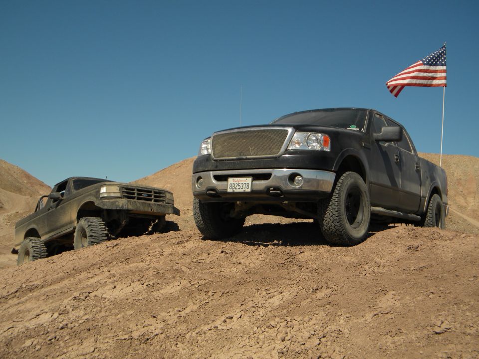 Black Ford F-150 with American flag and older brown truck on desert hill
