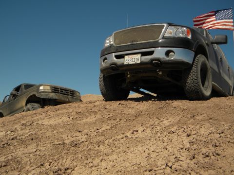 1992 Ford Bronco - A black Ford F-150 and a dirt-covered Ford Bronco are parked on a sandy hill under a clear blue sky.