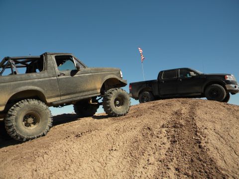 1992 Ford Bronco - Two trucks, a modified SUV and a pickup, navigate a dusty incline under a clear blue sky.