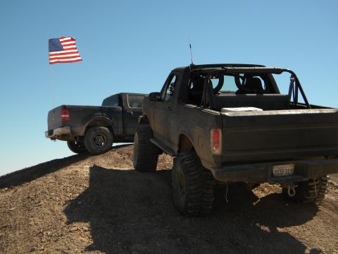 1992 Ford Bronco - Two modified trucks are parked on a dusty incline under a clear blue sky, with an American flag waving in the breeze.