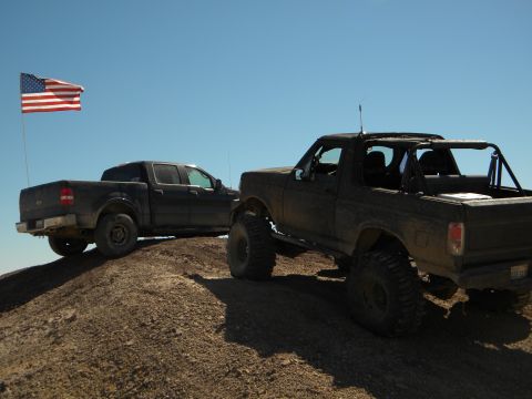 1992 Ford Bronco - Two modified trucks, including a 1992 Ford Bronco, sit atop a dirt mound under a clear blue sky with an American flag waving.