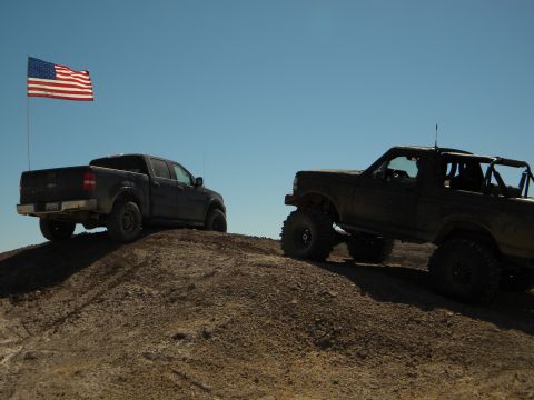 1992 Ford Bronco - A lifted 1992 Ford Bronco is seen off-roading next to a pickup truck under a clear blue sky.