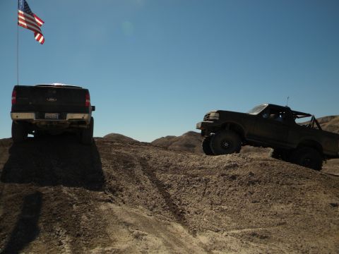 1992 Ford Bronco - Two modified trucks navigate a steep, dusty incline under a bright, cloudless sky.