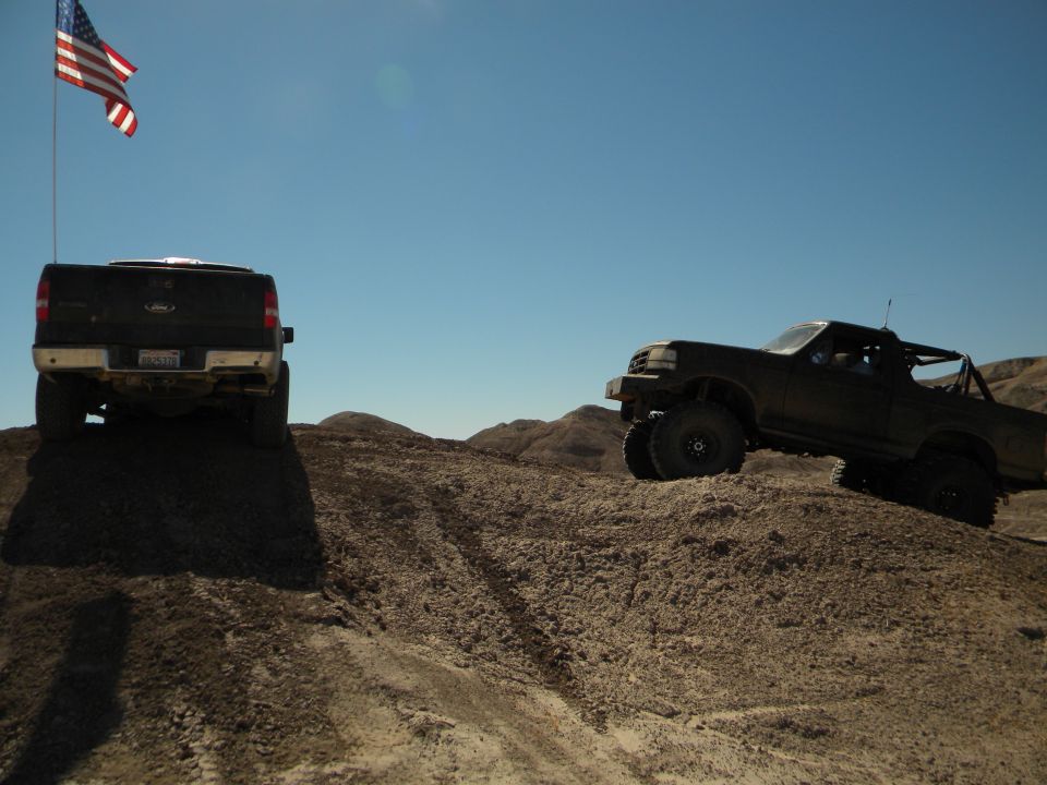 Two trucks are off-roading on a dirt hill under a clear blue sky with an American flag flying