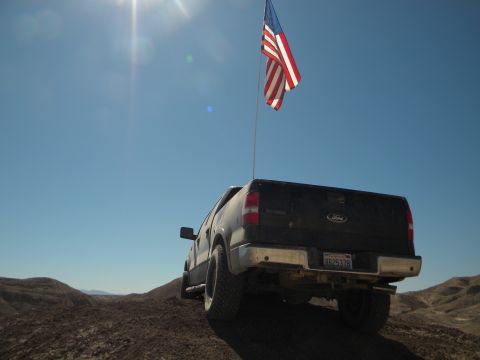 1992 Ford Bronco - A black Ford F-150 pickup truck flies an American flag while driving off-road on a sunny day.