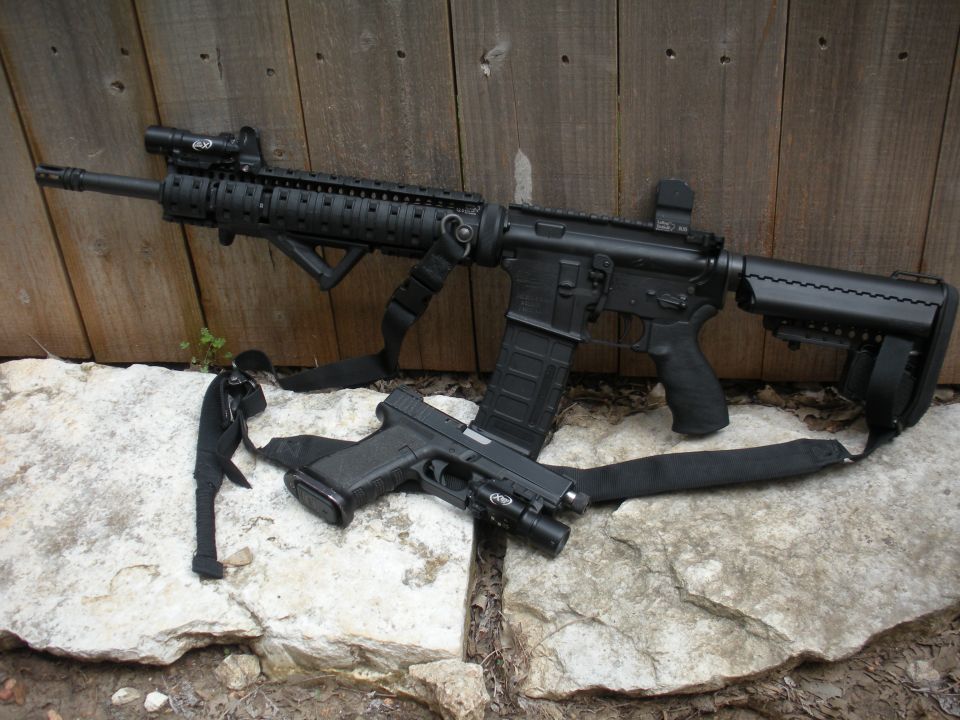 Black assault rifle and handgun resting on rocks against a wooden fence