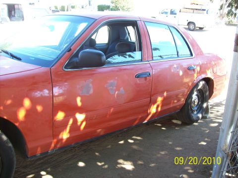 2002 Ford Crown Victoria - Side view of an orange 2002 Ford Crown Victoria sedan with visible paint damage and shadows from trees.