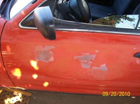 2002 Ford Crown Victoria - The side of a red 2002 Ford Crown Victoria shows significant paint damage and peeling, revealing primer underneath.