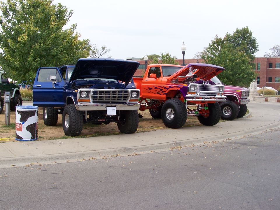 Three lifted Ford trucks parked side-by-side, hoods up. The front truck is blue, the middle is orange with flames, and the rear is purple