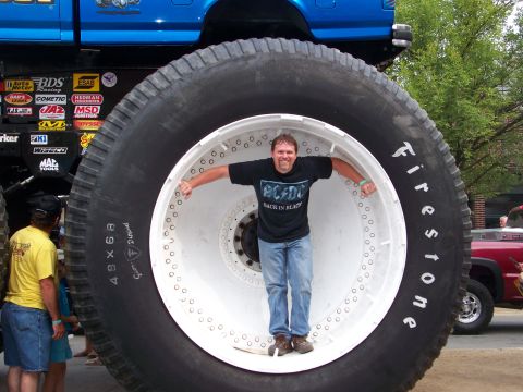 1989 Ford Bronco - Man poses inside a giant tire, with a monster truck and another vehicle visible.