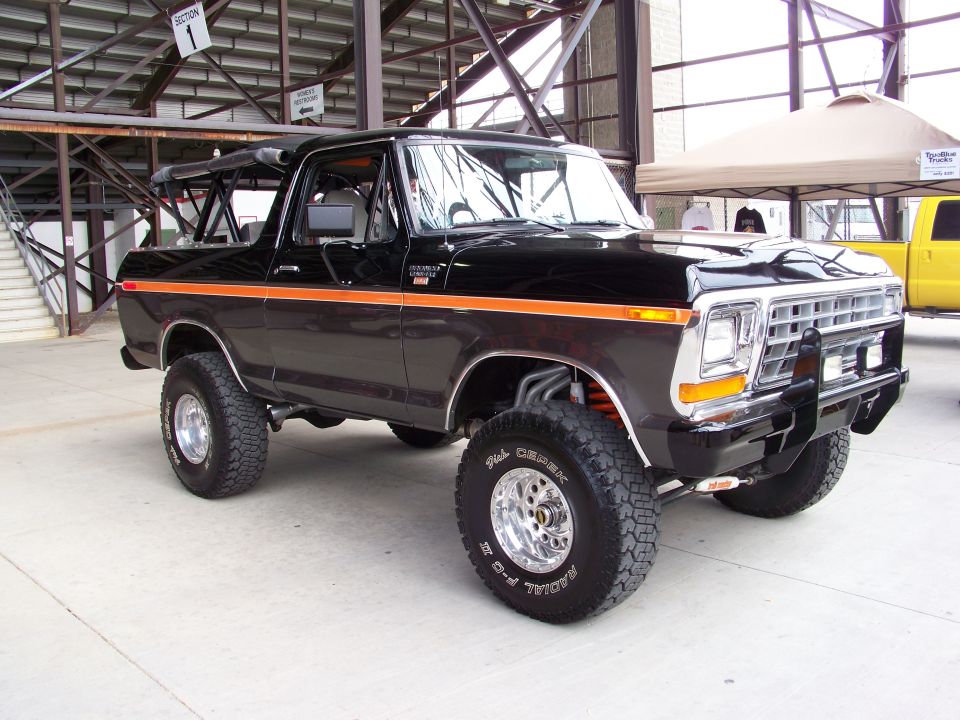 Black Ford Bronco with orange stripe, lifted suspension, and aftermarket wheels. Yellow truck visible behind a tent