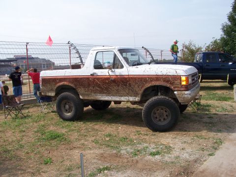 1989 Ford Bronco - A muddy Ford Bronco sits off-road, with a dark blue pickup truck visible behind it.