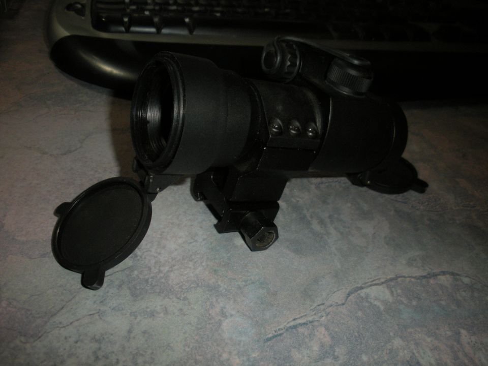 Black optical scope with lens cap open, mounted on a dark surface