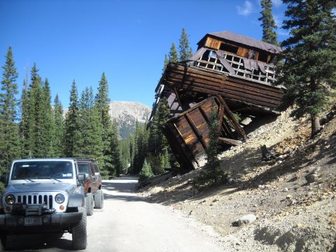 1986 Ford Bronco II - Off-road vehicles travel a dusty mountain path towards an old, unstable wooden structure.