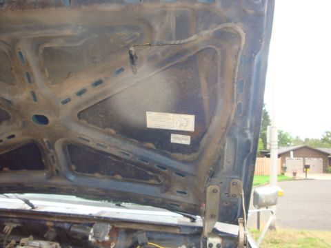 1989 Ford Bronco - The underside of a raised dark-blue truck hood, showing the ribbed stamped inner bracing, a hinge, and an emissions/vacuum-routing decal af…